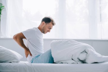 side view of young man in pajamas sitting on bed and suffering from back pain clipart