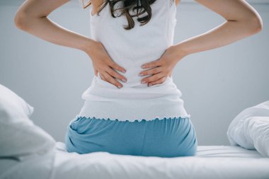 back view of young woman in pajamas suffering from back pain on bed