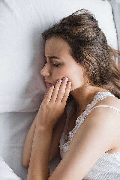 top view of young woman suffering from tooth pain while lying in bed 