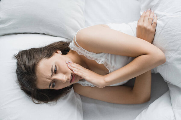 top view of young woman suffering from toothache while lying in bed 