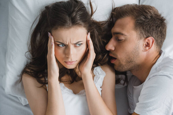 Top view of young woman with headache looking at husband snoring in bed
