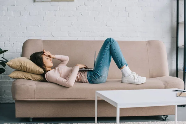自宅のソファの上に横たわっている間ノート パソコンを使用して頭痛を持つ若い女性 — ストック写真