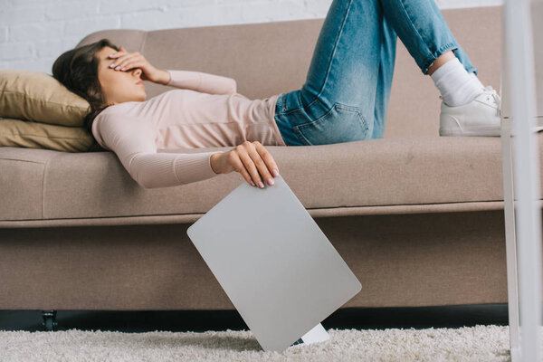 young woman with headache and pain in eyes lying on sofa and holding laptop