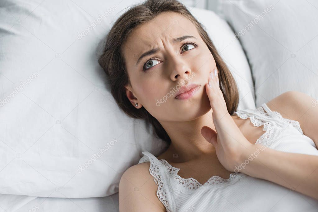 attractive young woman suffering from toothache and looking away while lying in bed 