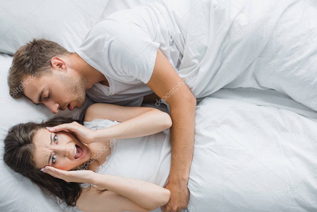 top view of young angry woman having headache and screaming while lying with snoring husband in bed 