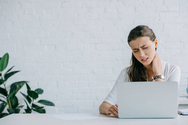 молодая деловая женщина, страдающая от боли в шее во время работы с ноутбуком
 