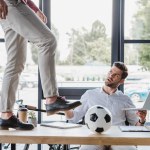 Recortado tiro de hombre pateando pelota de fútbol en la mesa mientras colega usando portátil en la oficina