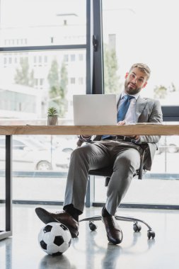 dizüstü bilgisayar kullanarak ve kamera Office gülümseyen futbol topu ile genç işadamı 