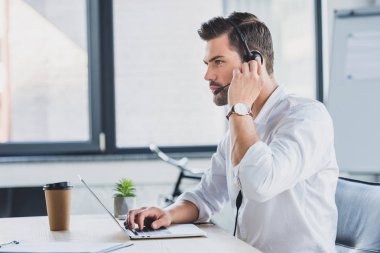 Genç çağrı merkezi operatörü dizüstü kullanarak ve uzak ofis seyir kulaklık yan görünüm