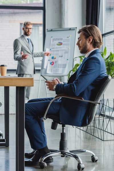 Jungunternehmer Bedient Smartphone Während Kollege Auf Whiteboard Zeigt — kostenloses Stockfoto