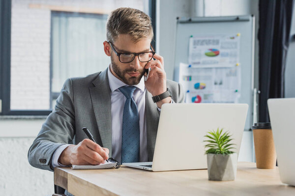serious businessman in eyeglasses talking by smartphone, taking notes and using laptop in office
