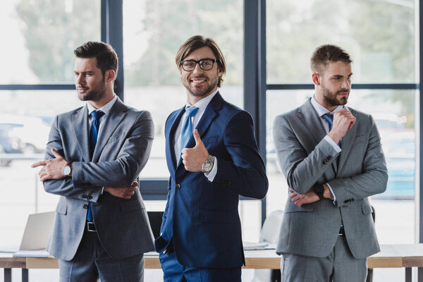 handsome young businessman in eyeglasses showing thumb up and smiling at camera while standing with confident colleagues in office