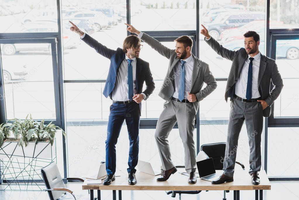 three happy young businessmen dancing on table in office 