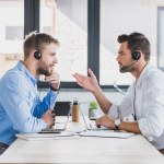Zijaanzicht van jonge call center exploitanten in hoofdtelefoons bespreken en kijken naar elkaar tijdens het gebruik van laptops in kantoor