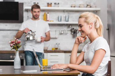 gülümseyen genç kadın kulaklık dizüstü erkek Kupası ve su ısıtıcısı mutfakta elinde iken kullanma 