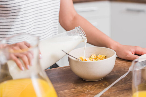 cropped image of girl pouring milk from bottle into plate with cornflakes during breakfast in kitchen