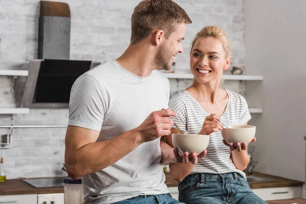 smiling couple holding plates and spoons with corn flakes and looking at each other in kitchen