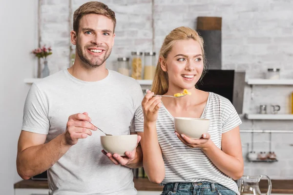 happy couple holding plates and spoons with corn flakes in kitchen