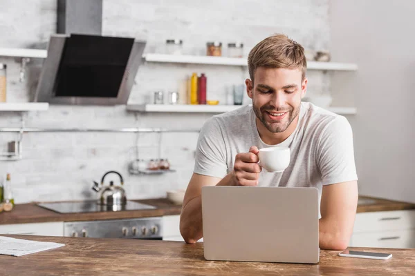 Bonito Sorrindo Homem Segurando Xícara Café Olhando Para Laptop Cozinha — Fotografia de Stock