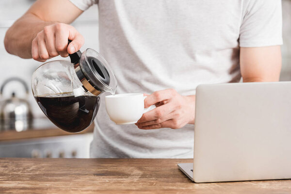 обрезанное изображение человека, наливающего кофе в чашку возле ноутбука на кухне
