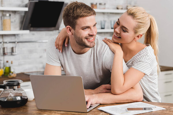 smiling couple sitting in kitchen with laptop