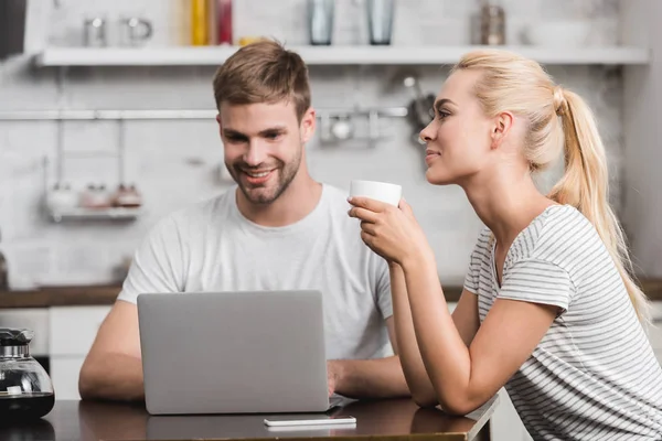 微笑的年轻妇女拿着杯咖啡 而快乐的男朋友使用膝上型电脑在家中 — 免费的图库照片