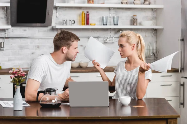 情感的年轻夫妇争论和看着对方 而使用笔记本电脑 并在家里拿着报纸 — 图库照片