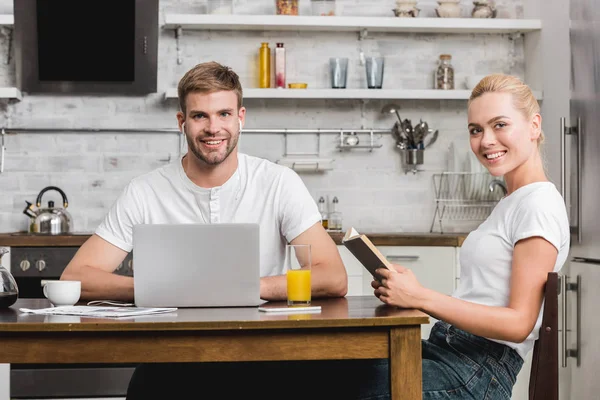 快乐的年轻夫妇与笔记本电脑和书坐在厨房的桌子和微笑的相机 — 图库照片