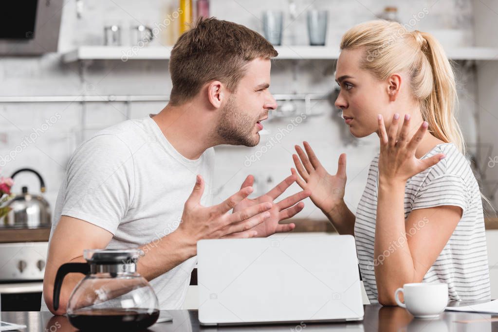 side view of emotional young couple arguing and looking at each other in kitchen, relationship difficulties concept 