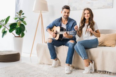Akustik gitar ile kanepede oturan ve evde kameraya bakarak genç gülümseyen çift 