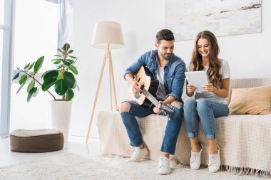 smiling couple tuning acoustic guitar with digital tablet on sofa at home clipart