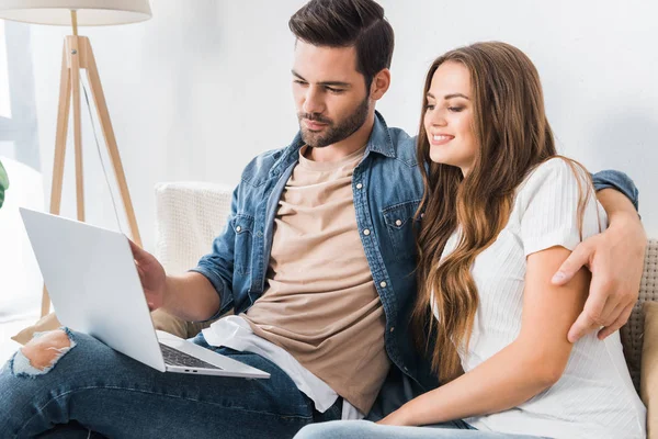 自宅のソファの上のガール フレンドをノート パソコンの画面に指で指しているハンサムな男 — ストック写真