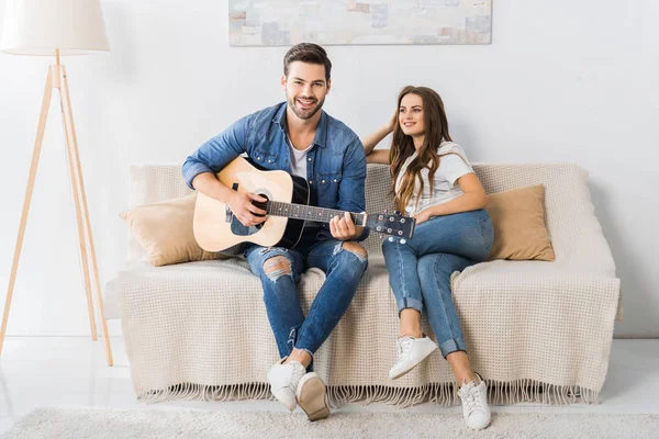 Jovem Tocando Guitarra Acústica Enquanto Sua Namorada Sentada Perto Sofá — Fotografia de Stock Grátis