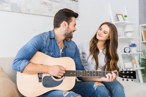 happy young man playing on acoustic guitar and looking at his girlfriend on sofa at home 