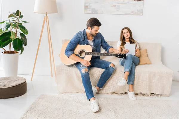 デジタル タブレットの自宅のソファの上でアコースティック ギターをチューニングする若いカップル — ストック写真