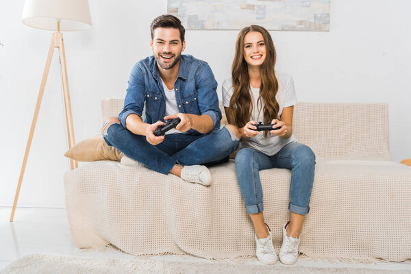 улыбающаяся молодая пара играет в видеоигры на диване дома
 
