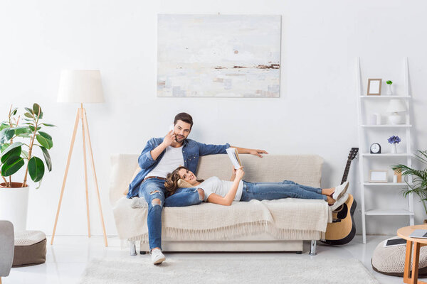 young man talking on smartphone while his girlfriend reading book on sofa at home 