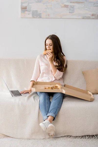 自宅のソファーで休んでいる間ノート パソコンを使用してピザを持つ若い女性 — ストック写真