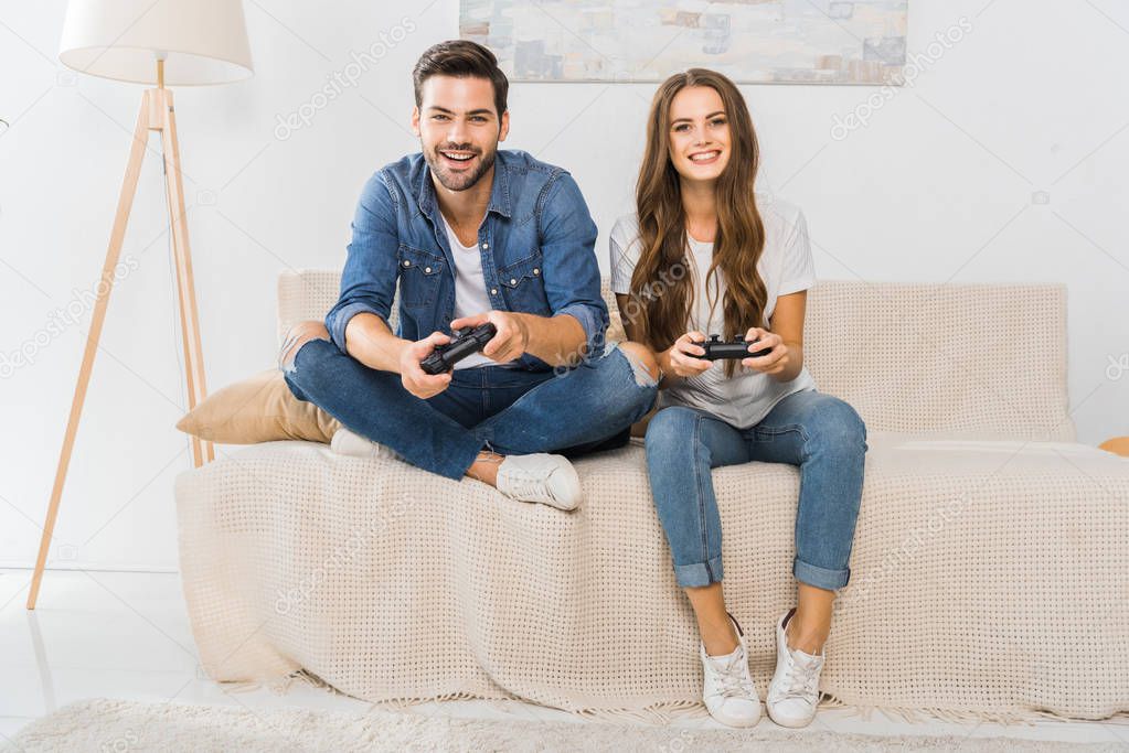 smiling young couple playing video game by joysticks on sofa at home 