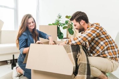 smiling couple unpacking cardboard boxes together at new home, moving home concept