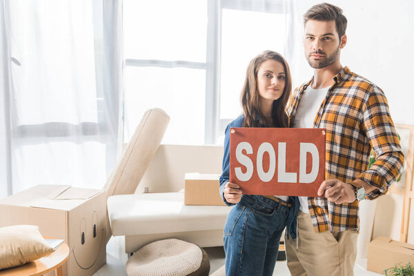 portrait of young couple holding sold red card at home with cardboard boxes