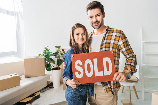 portrait of smiling couple holding sold red card at home with cardboard boxes