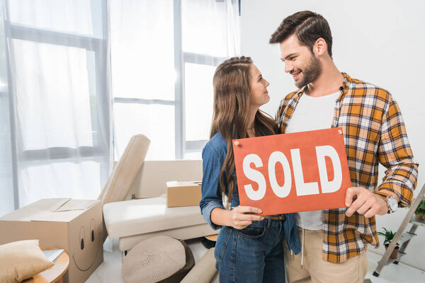 portrait of couple holding sold red card at home with cardboard boxes