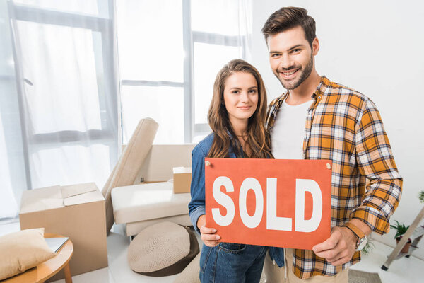 portrait of smiling young couple holding sold red card at home with cardboard boxes