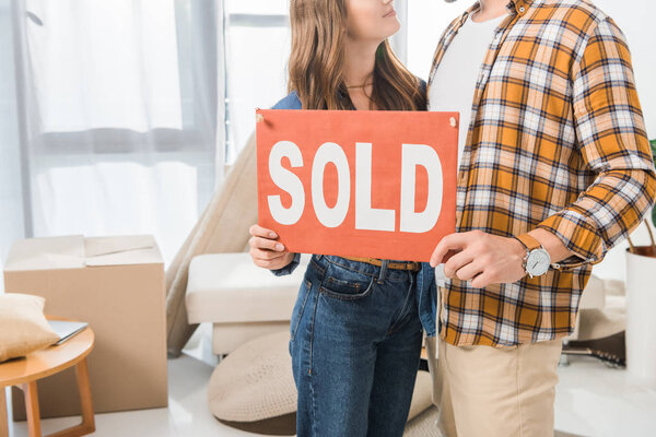 partial view of couple holding sold red card at home with cardboard boxes