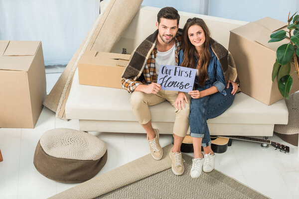high angle view of young couple in blanket with our first house card sitting on sofa, moving home concept
