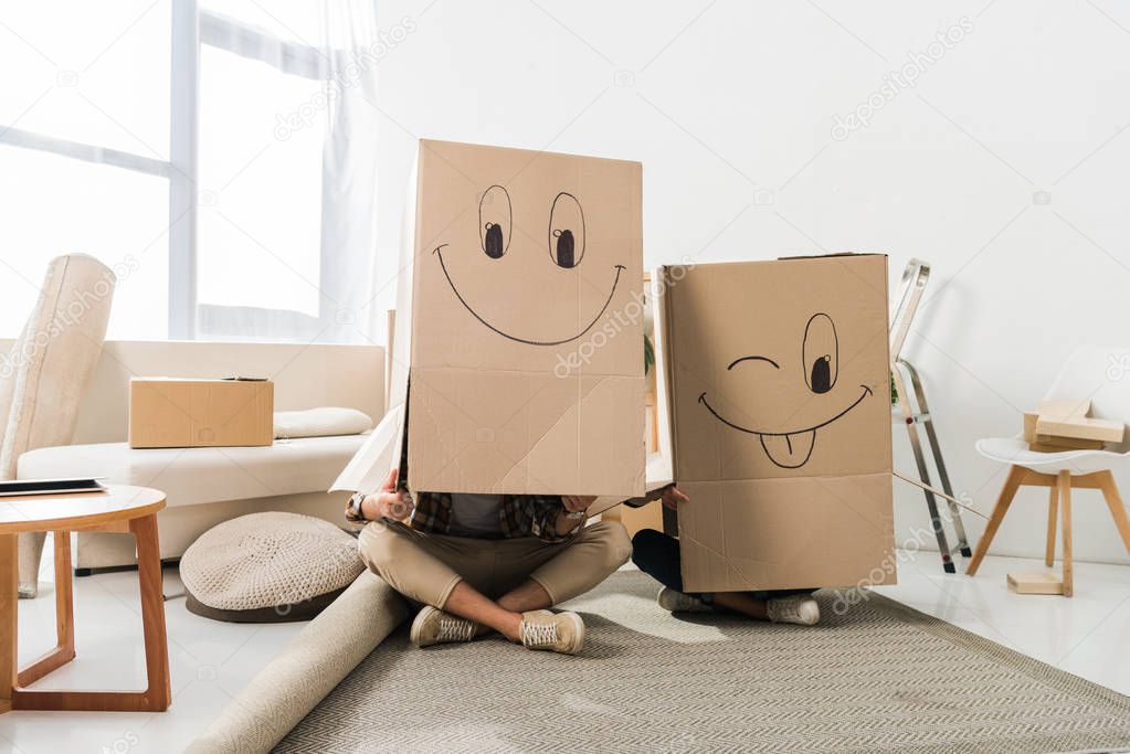 obscured view of couple with cardboard boxes on heads sitting on floor at new house, moving home concept