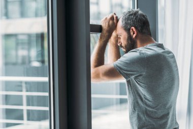 depressed bearded man leaning at window and looking through it 