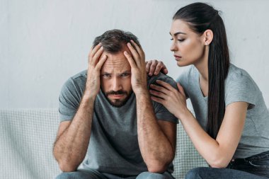 young woman supporting depressed frustrated boyfriend sitting on couch and looking at camera clipart