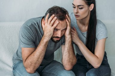 young woman supporting depressed frustrated boyfriend sitting on couch   clipart